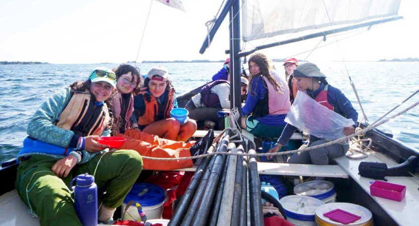 A group of students wearing life jackets smile while sitting in a sailboat